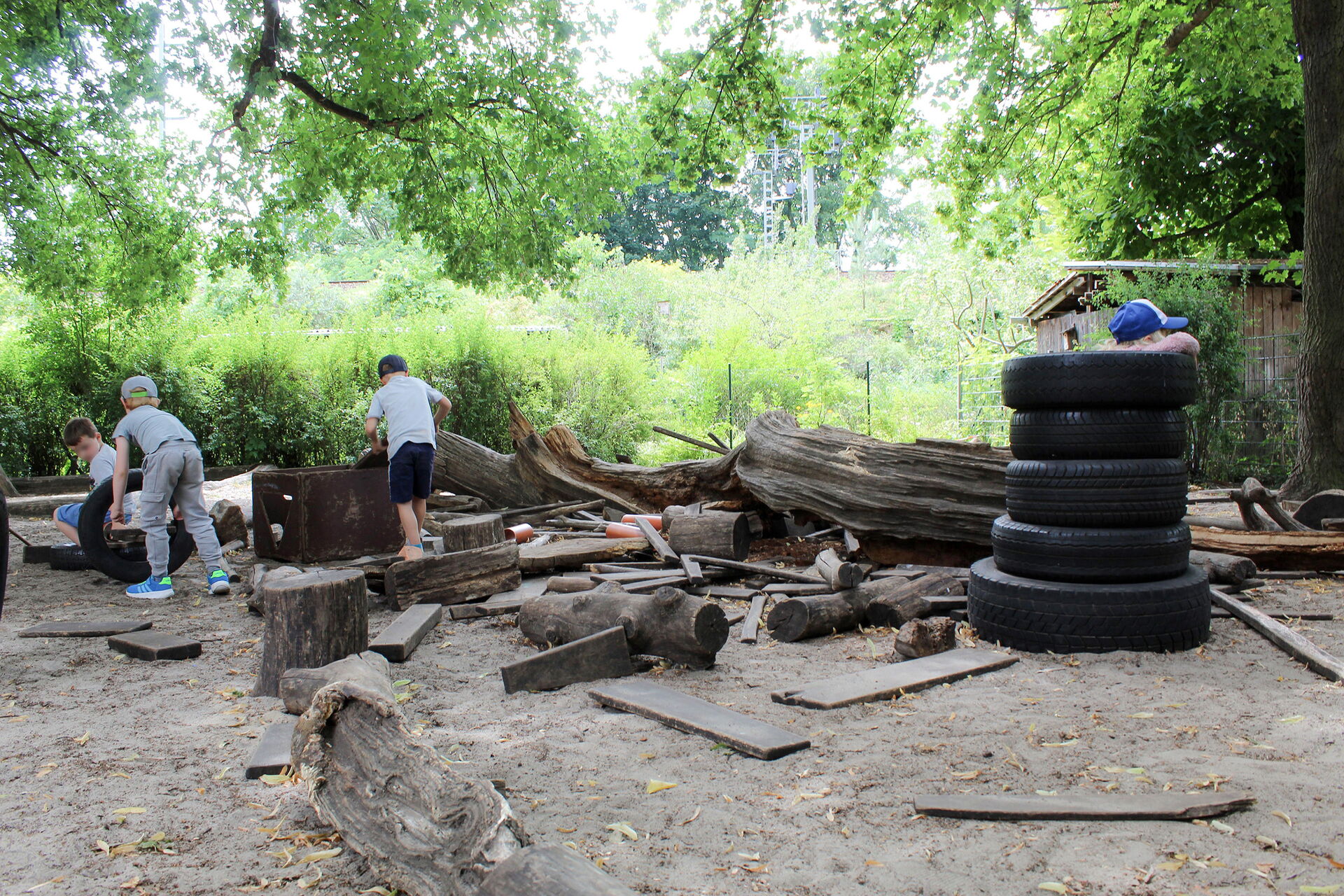 Holz-Baustelle im Garten. Reifen sind aufgestapelt und Holzstücke, Holzstämme liegen rum. 4 Kinder spielen dort.