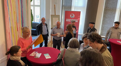 Ein Raum mit roten Stehtischen. Im Raum 12 Menschen, die beieinanderstehen, sich unterhalten. An einem Tisch steht Frau Schweers mit einem Hefter mit Papieren.