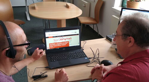 Zwei Männer schauen auf einen Laptopmonitor. Ein Mann trägt Kopfhörer.