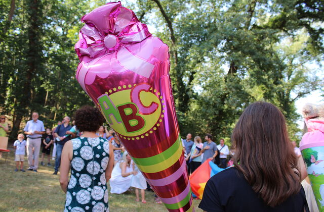 Großer Luftballon in Form einer Zuckertüte pink mit A-B-C Bild. Einige Menschen draußen auf einer Wiese mit Bäumen.