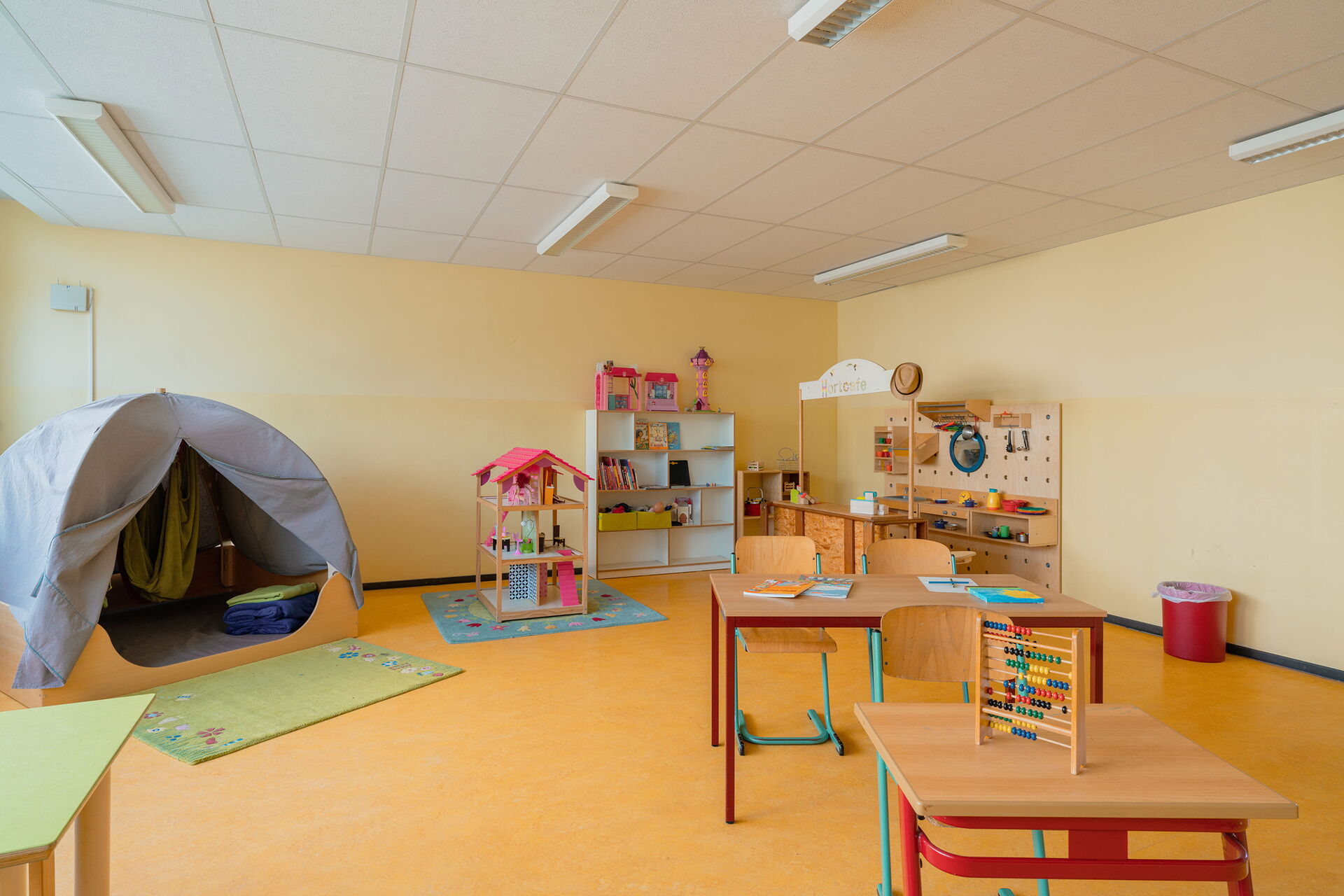 Spielzimmer, auf einem kleinen Tisch steht ein Rechenschieber. Ein Puppenhaus steht auf einem Kinderteppich, ein Regal an der Wand mit Spielzeug: