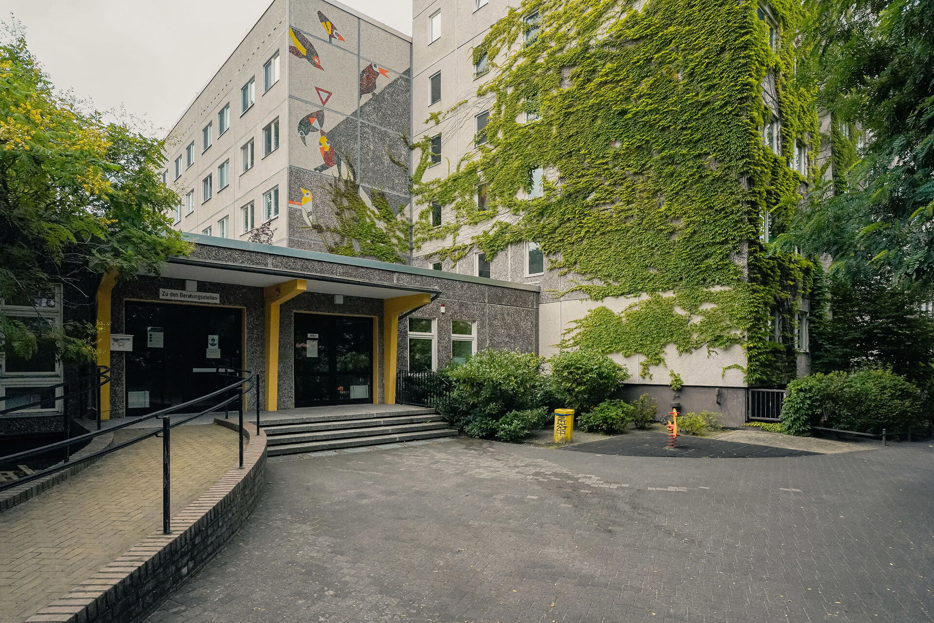 Eingang des Gebäudekomplex. Das Gebäude ist mit Efeu bewachsen. Zum Eingang Stufen und links eine geschwungene Rampe.