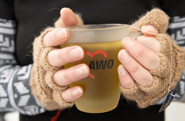 Teetasse mit AWO-Logo. Tasse wird mit zwei Händen in fingerlosen braunen Handschuhen gehalten.