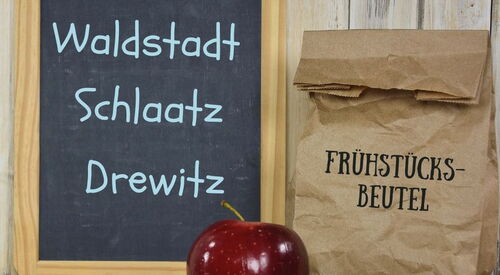 Eine Tafel, an der mit blauer Kreide geschrieben steht: Ab 9.11. Waldstadt Schlaatz Drewitz - Das Kreidestück liegt vor der Tafel sowie ein roter Apfel. Daneben ist eine gefüllte Papiertüte darauf steht: Frühstücksbeutel
