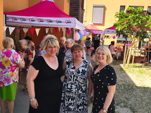 Gruppenfoto: Draußen, 3 Frauen in Sommerkleidern stehen vor bunt geschmückten Pavillons.