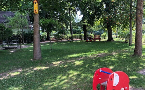 Weitläufiger Spielplatz, Rasen schattig durch einige Bäume. Vorne ein kleiner roter Elefant zum raufsetzen und reiten für kleine Kinder.