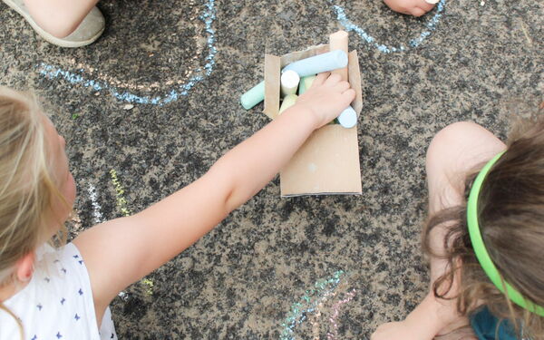 4 kleine Kinder sitzen / hocken am Boden auf Stein. In der Mitte ein kleiner Karton mit Kreide. Sie malen mit Kreide.