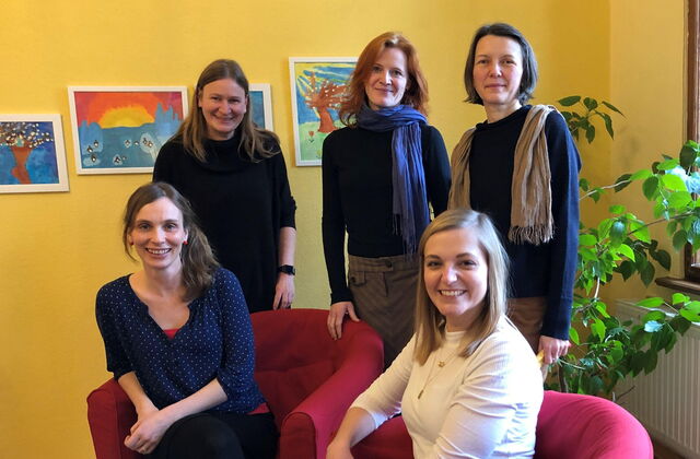Fünf Frauen in einem Raum mit gelben Wänden und Kinderbildern an der Wand. Zwei Frauen sitzen in roten Sesseln, drei stehen dahinter. Alle lächeln freundlich in die Kamera.