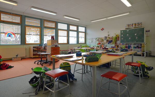 Lernzimmers. großer Tisch in der Mitte des Raumes, darauf Federtaschen, Blöcke und Hefter. Um den Tisch stehen Hocker und Schulranzen.Vorne eine grüne Schultafel.