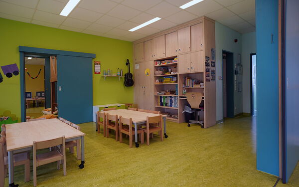 Ein Kindergartenraum mit Tischen und Stühlen, einer Wand und Fußboden in grüner Farbe. Regale und Schränke.