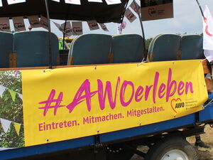 Ein Traktor Anhänger mit #AWO erleben Banner und AWO Wimpeln. Hinten schiebt ihn ein Mann im blauen T-Shirt an.