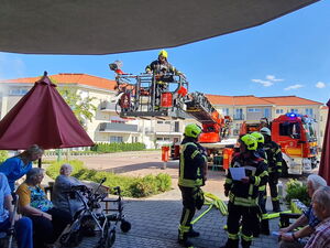 Feuerwehrauto mit Kran und viele Feuerwehrleute. Senioren sitzen im Schatten vor den Gebäude.