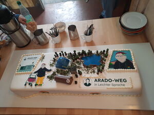 Eine rechteckige Torte mit weißem Zuckerguss ist verziert mit Figuren, Pyramide, Schokoriegel und Flächen mit schwarzer Schrift: Arado-Weg in Leichter Sprache