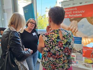 Drei Frauen stehen am Stand vom Netzwerk Arbeit Inklusiv für Potsdam der AWO Potsdam. Die Frauen sind in einem Gespräch/Austausch. Info-Material liegt auf dem Stand-Tisch.