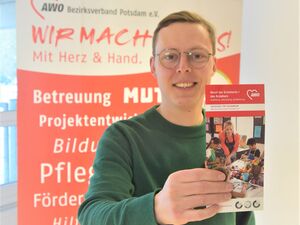 Junger Mann mit einem Flyer der Einrichtung lächelt in die Kamera. Er hat kurze braune Haare, eine Brille mit silbernen Rahmen und einen grünen Pullover.