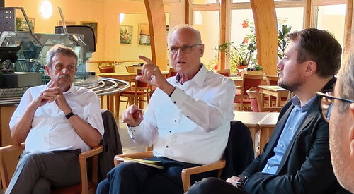 In einer Kantine/Cafeteria. 3 Männer sitzen im Halbkreis. Mann mit Brille und weißem langärmligen Hemd spricht. Es benutzt auch die Hände um etwas zu verdeutlichen.
