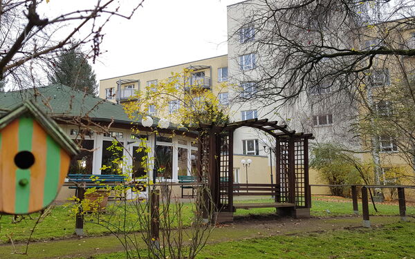 Parkanlage mit kleinem Häuschen mit grünem Dach. Vorne links ist ein kleines Vogelhäuschen. Im Hintergrund ein großes Wohnhaus.