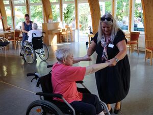 In einem großen Speiseraum. Zwei alte Frauen in Schieberollstuhlen tanzen mit zwei Frauen, die sie an den Händen fassen und hin und her ziehen/schieben.