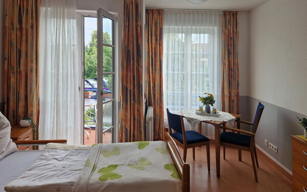 Zimmer mit Bett, Tisch und zwei Stühlen, die Balkontür ist offen. Blick zum Balkon und Fenster.