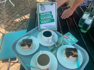 Ein Tablett mit Kaffee und Kuchen und einem Flyer von Kultur für Jeden. auf einem Tisch. Auf dem Tisch stehen zwei Wasserflaschen und ein kleiner Aufsteller mit kleinem Plakat. Text: Keffee, Kuchen,, Kunst! Herzlich willkommen im museums Café.