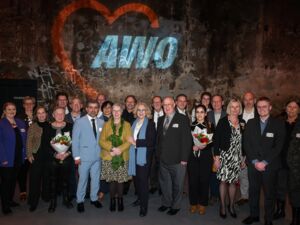Gruppenfoto. Viele Menschen vor einer Wand mit dem AWO-Logo. Alle sind festlich / feierlich gekleidet. Zwei Frauen haben Blumensträuße in der Hand.