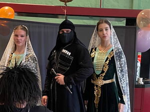 Vier Personen in schwarzen Kostümen mit weißen Schleiern und Verzierungen, eine davon kniet mit ausgebreiteten Armen im Vordergrund.