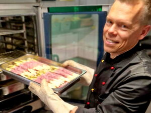 Koch lächelt in die Kamera. Er hat ein Blech mit Spargel in Schinken gerollt in behandschuten Händen und schiebt es in den Ofen.