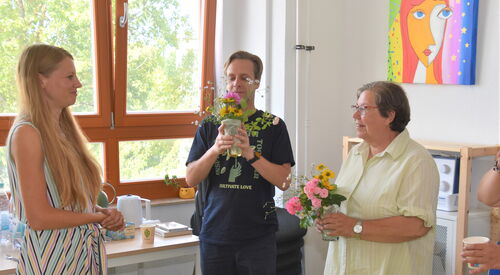 Beide Künstler stehen nebenienander und halten kleine Blumensträuße im Wasserglas in der Hand. Jüngerer Mann in blauem T-Shirt und kurze Hose. Ältere Frau, kurze graue Haare und Brille mit heller Bluse. Freu mit langen blonden Haaren steht ihnen gegenüber