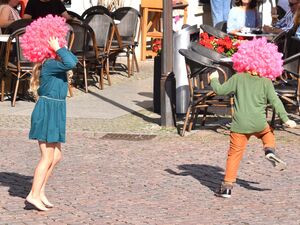 Draußen, Sonne, zwei Kinder mit pinken Clowns-Perücken tanzen wild herum. Ein Mädchen, barfuß, mit Kleid und langen blonden Haaren. Das andere Kind von hinten, grüner Pulli, orangene Hose.