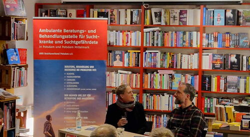 Eine Frau und ein Mann sitzen an einem Tisch und sehen sich an. Neben dem Tisch ein Roll-Up der AWO Suchtberatung.       Hinter ihnen große Bücherregale. Publikum sitzt im Raum.