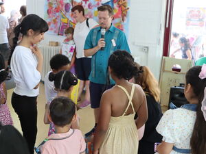 Mann in blauen Hemd spricht in ein Mikrofon und wird dabei von Kindern umringt.