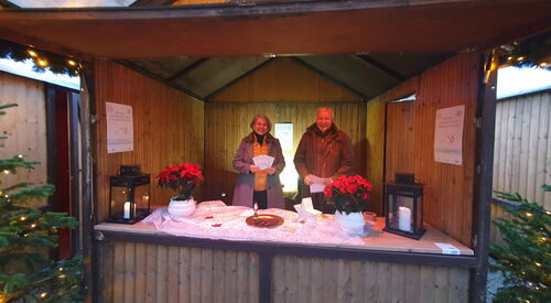 Weihnachtlicher Stand aus Holz mit Tannenbäumen rechts und links. Zwei Weihnachtssterne-Pflanzen und eine Frau und ein Mann im Stand.