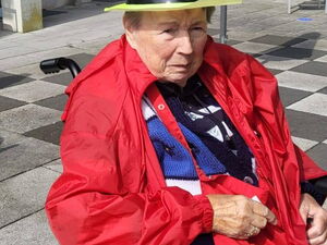 Seniorin im Rollstuhl auf sonniger Terrasse. Sie trägt rote Kleidung und einen gelben Hut.