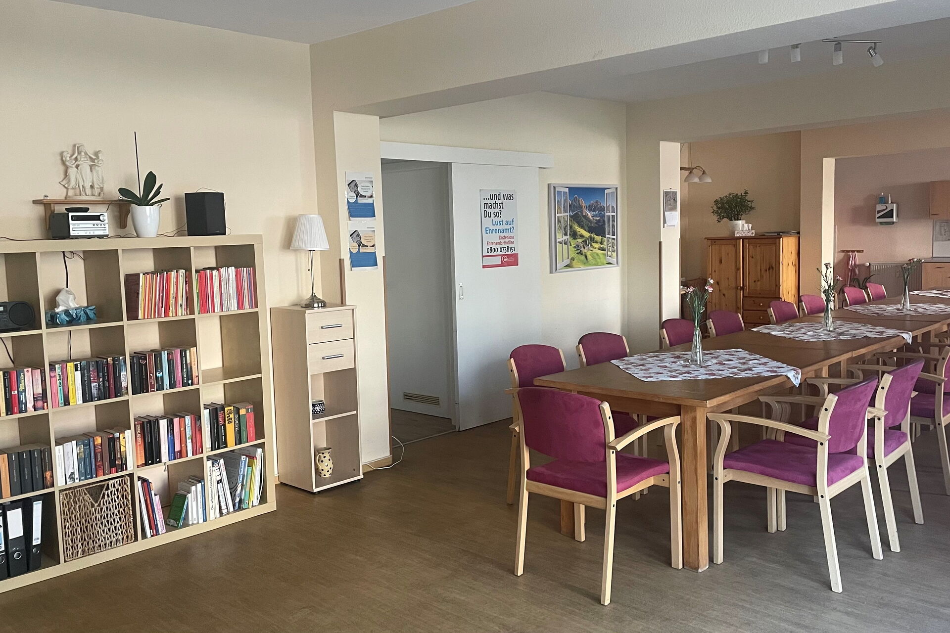 am linken Bildrand steht ein ausreichend gefülltes Bücherregal, darauf steht eine Musikanlage. rechts davon steht in der Mitte des großen Raumes eine lange Tischtafel mit Stühlen.