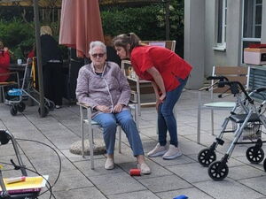 Büchsenwerfen. Seniorin sitzt auf Stuhl auf der Terrasse. Betreuerin steht neben ihr. Auf dem Boden liegen Dosen.