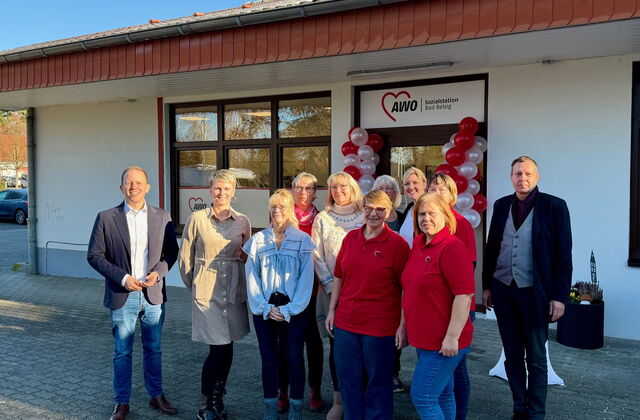 11 Personen, zwei Männer links und rechts außen stehen vor einem weißen Gebäude. Der Eingang ist mit roten und weisen Luftballons geschmückt.