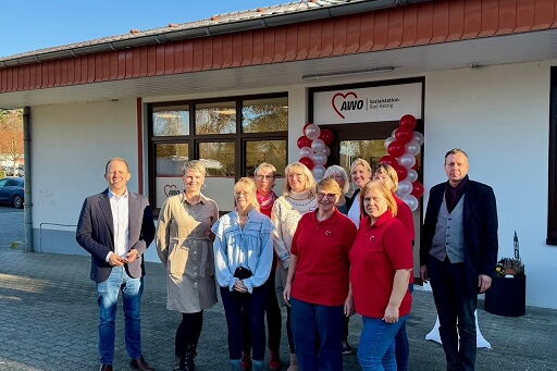 im Vordergrund Personengruppe mit 11 Personen, im Hintergrund Gebäude mit roten und weißen Luftballons rechts und links des Haupteinganges, strahlender Sonnenschein und blauer Himmel