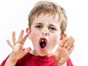 Frontal, ein Junge drückt sich an einer Scheibe die Nase platt und zieht eine Grimasse. Die Haare sind hellbraun und verwuschelt.