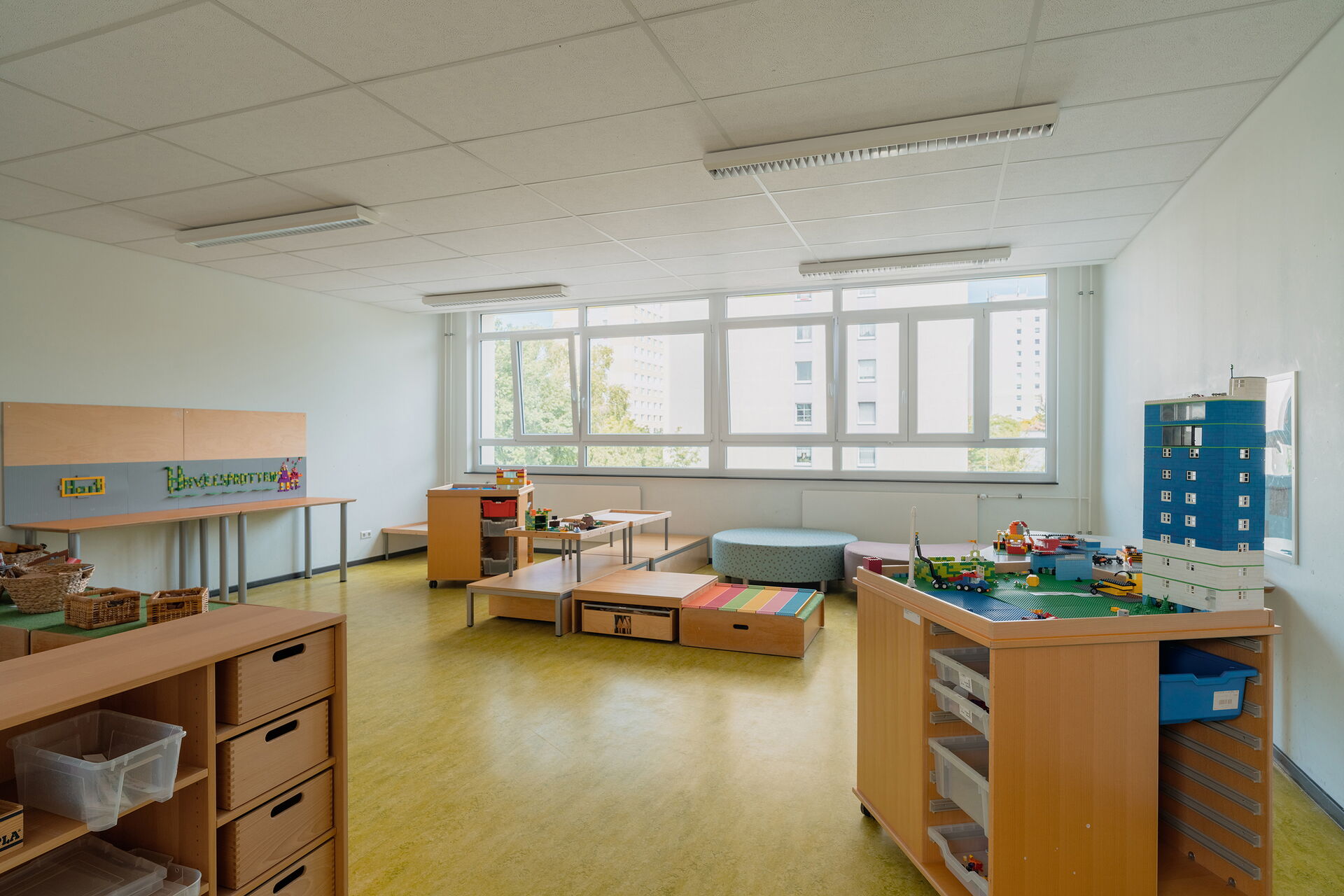 Spielzimmer. Viel Platz, große Fenster. Sitzmöbel bei den Fenstern. Lego Tisch mit Hochhaus bebaut.