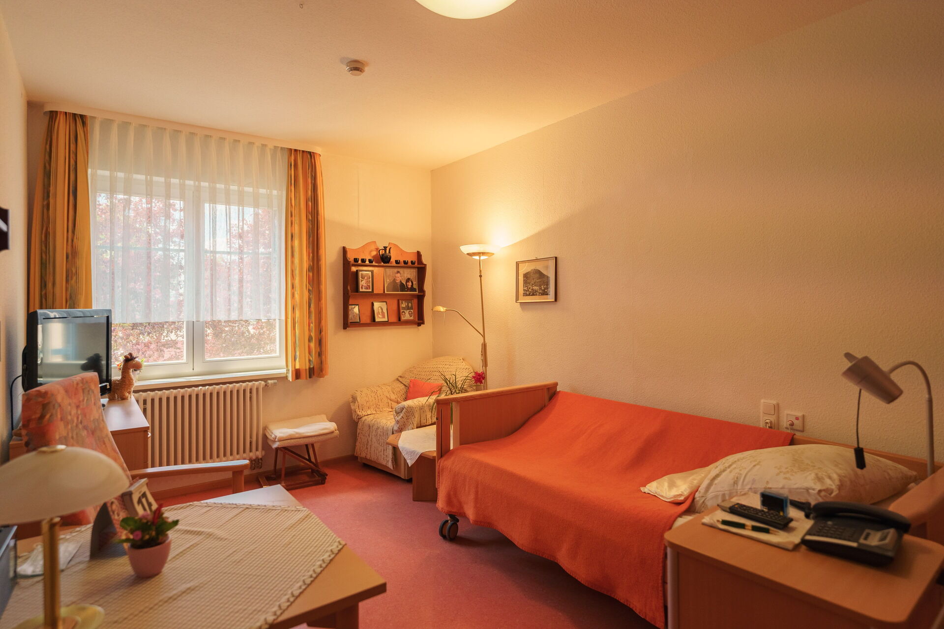 Zimmer mit Bett, Sessel in der einen Ecke, Fernseher in der Ecke gegenüber und kleinem Tisch.