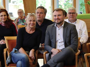 6 Personen sitzen auf Stühlen. Frontal. Ganz vorne eine blonde Frau mit Brille auf den Kopf geschoben, daneben ein Mann im grauen Anzug und weißen Hemd. Beide lächeln leicht in die die Kamera.