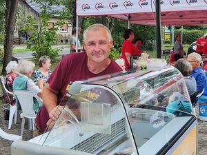 Ein Mann steht an einer Eistheke. Er hat sich daran angelehnt und lächelt in die Kamera. Er trägt ein weinrotes T-Shirt mit gelben Aufdruck. Hinter ihm ist ein AWO Pavillon und ältere Menschen sitzen auf Gartenstühlen.