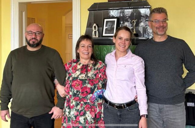 Raum mit gelben Wänden. 4 Personen stehen nebeneinander und haben sich unterhakt, 2 Männer rahmen 2 Frauen ein. Die Männer tragen Brille, Bart und dunkle Pullover. Frau trägt ein Blumenkleid, die Andere trägt eine langärmlige hellrosa Bluse. Alle lächeln