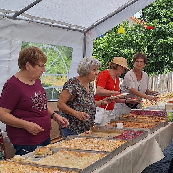 Kuchenbüfett. 7 große Blechkuchen auf einen langen Tisch. 4 Frauen stehen hinter dem Tisch um Kuchen aufzutun.