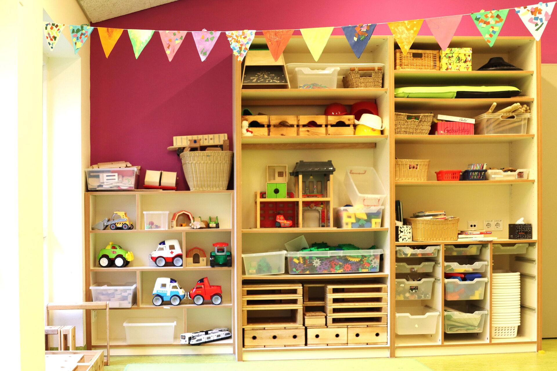 Heller Raum, großes Regal steht an der Wand. Körbe und Aufbewahrungskisten im Regal. links daneben in der Ecke ein kleines Regal mit Spielzeug, Autos, Bagger