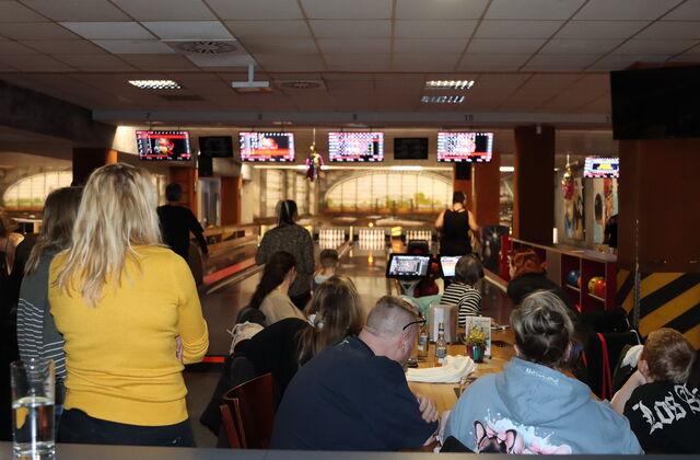 In einem Bowlingcenter. Im Vordergrund Menschen an Tischen mit Getränken. Dahinter die Bowlingbahnen.