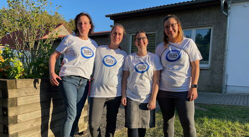Draußen, Sonne scheint. 4 Frauen stehen nebeneinander und lächeln in die Kamera. Sie tragen einheitliche weiße T-Shirts mit Aufdruck: Blauer Kreis: Kinder stark machen steht drauf.