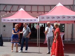 Frau in langem roten Sommerkleid und Mann in weißem Poloshirt und grauer Hose mit Mikros in der Hand. Die Frau blickt zu zwei anderen Menschen. Frau mit roten langen Haaren und blauen kurzen Kleis und Mann im grauen Hemd und Jeans wirken froh.