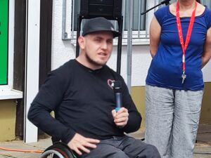 Mann im Rollstuhl mit Mikrofon in der linken Hand, er trägt ein schwarzes Base Cape und einen schwarzen Pulli.