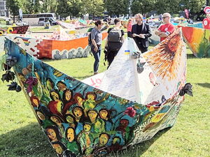 Viele bunt bemalte große Papierboote auf einer Wiese. Dazwischen vereinzelt Menschen.